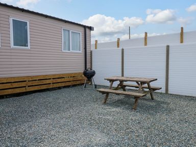 An outdoor area with gravel ground a wooden picnic table a barbecue grill next to a mobile home and white fencing at Trewan Lodge in Bryngwran near Rhosneigr