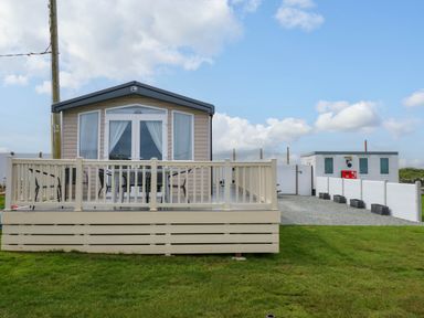 A small beige cabin with a front deck and outdoor furniture on a grassy plot at Trewan Lodge Bryngwran near Rhosneigr
