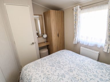 A bedroom with a bed covered in floral bedding a wooden wardrobe a small vanity with a mirror a stool a window with curtains and a radiator at Wansbeck View 2 in Ashington