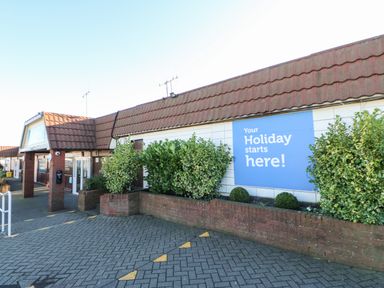 The exterior of a building with a tiled roof and a blue sign that says your holiday starts here with plants and a brick planter bed at Wansbeck View 2 in Ashington