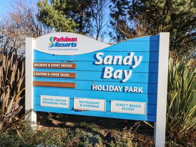 A blue sign for Sandy Bay Holiday Park with information about heated pool restaurant and direct beach access at Parkdean Resorts
