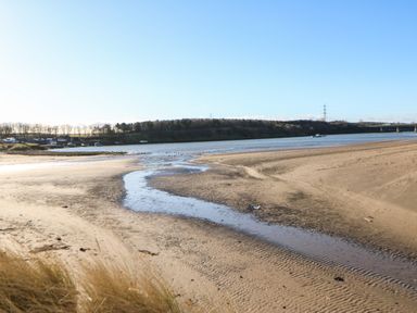 A sandy riverbank with a small stream flowing into the river and boats docked near trees at Wansbeck View 2 in Ashington