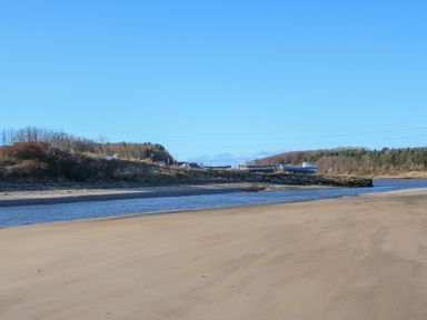 A sandy beach with a small river or inlet and boats on a stone jetty with trees in the background at Wansbeck View 2 in Ashington