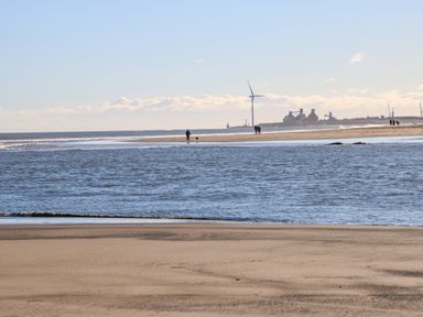 A sandy beach with water and people walking dogs near a wind turbine and buildings at Wansbeck View 2 in Ashington