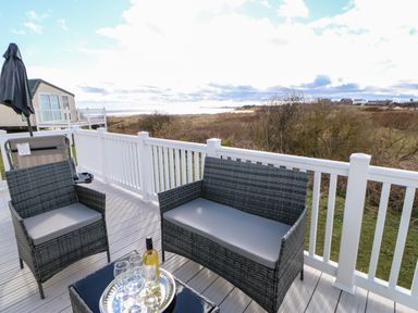 An outdoor deck with wicker chairs a table with a wine bottle and glasses and a view of a grassy area and water at Wansbeck View 2 in Ashington