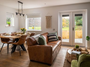 A dining area with a wooden table and brown chairs next to a living room with a brown leather sofa and a round wicker coffee table at Sygun in Beddgelert