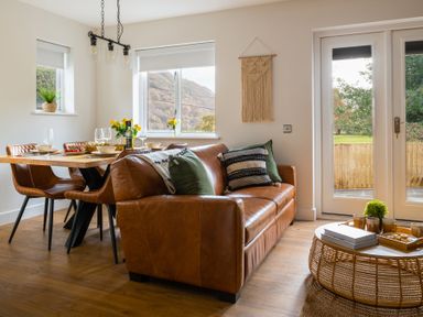 A dining area with a wooden table, four chairs, a brown leather sofa, round coffee table with cups and books at Sygun in Beddgelert