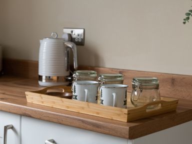 A kitchen counter with a wooden tray holding two mugs and three glass jars next to a white electric kettle at Sygun in Beddgelert