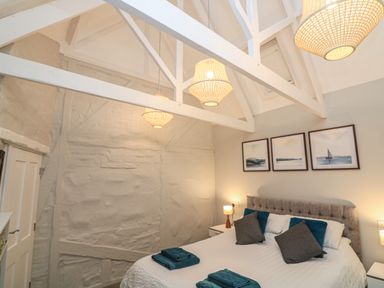 A bedroom with a white bed, gray headboard, pillows, bedside tables with lamps, framed artworks on the wall and exposed white ceiling beams at Queens View in Dartmouth