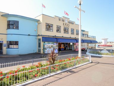 Exterior of a building with blue and beige walls flags on the roof and a fenced garden with flowers in front at Anchors Away in Weymouth