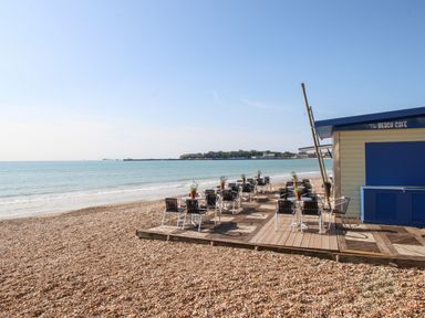 Outdoor seating area with tables and chairs on a wooden deck next to a beach cafe on a pebble beach by the sea at Anchors Away in Weymouth