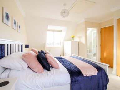 A bedroom with a bed with pillows and throw blankets a white dresser with flowers a window and a door leading to a bathroom at Anchors Away in Weymouth