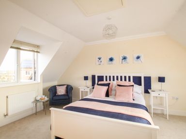 A bedroom with a white bed and pink and navy pillows two white nightstands with navy lamps a blue armchair by a window with blinds at Anchors Away in Weymouth