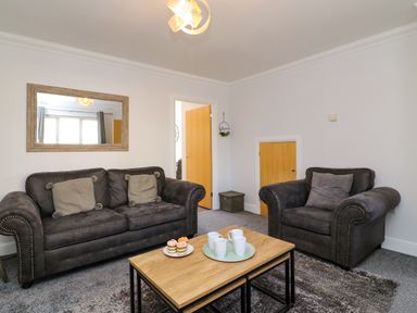 A living room with two dark sofas a wooden coffee table with cups and cupcakes a mirror on the wall and an open door at Anchors Away in Weymouth