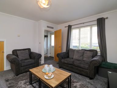 A living room with a sofa and armchair a coffee table with mugs and cupcakes a large window with curtains at Anchors Away in Weymouth