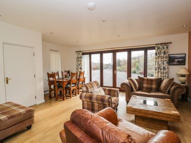 A living room with leather and plaid sofas a wooden coffee table and a dining table with chairs near large glass doors at The Lodge in Dol-y-Gaer near Pontsticill