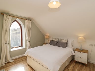 A bedroom with a bed pillows bedside tables lamps arched window with curtains and wooden floor at The Lodge in Dol-y-Gaer near Pontsticill
