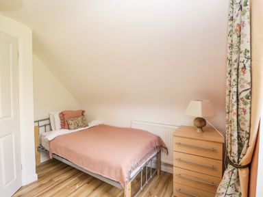 A small bedroom with a metal frame bed covered by a pink blanket and pillows next to a wooden chest of drawers with a lamp and floral curtains at The Lodge in Dol-y-Gaer near Pontsticill