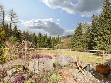 An outdoor garden area with rocks purple flowers wooden chairs and a metal fire pit surrounded by trees and fenced grass at The Lodge in Dol-y-Gaer near Pontsticill