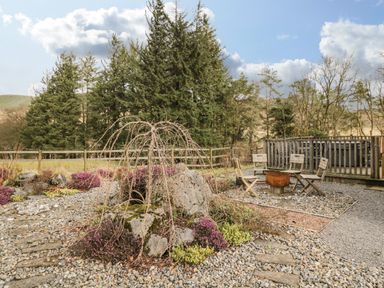 An outdoor seating area with wooden chairs around a fire pit and a hot tub on a deck in a garden with trees at The Lodge in Dol-y-Gaer near Pontsticill