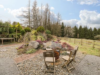 An outdoor seating area with four wooden chairs around a fire pit on a gravel and pebble surface near a garden and wooden fence at The Lodge in Dol-y-Gaer near Pontsticill