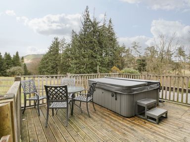 An outdoor wooden deck with a round glass table and four metal chairs next to a covered hot tub and surrounded by trees at The Lodge in Dol-y-Gaer near Pontsticill