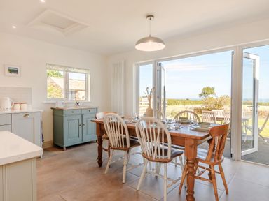 A dining area with a wooden table set with plates and bowls and white chairs with a large glass door opening to an outdoor patio at Morlan Cottage in Newport Pembrokeshire