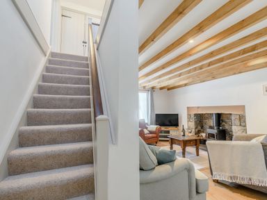 A carpeted staircase with wooden handrail next to a living room with exposed wooden ceiling beams stone fireplace and seating at Morlan Cottage in Newport Pembrokeshire