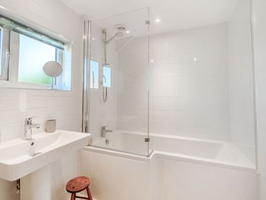 A bathroom with a sink, a stool, a window with a fish-patterned blind, and a bathtub with a shower and glass screen at Morlan Cottage Newport Pembrokeshire