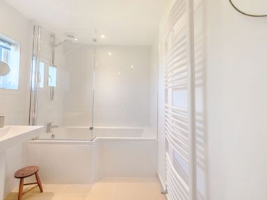 A bathroom with a bathtub shower combo a glass shower screen a white sink a small wooden stool and a white heated towel rail at Morlan Cottage in Newport Pembrokeshire