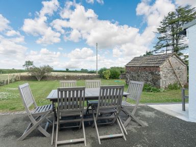 An outdoor patio with wooden table and chairs overlooking a grassy yard and a small stone building at Morlan Cottage in Newport Pembrokeshire