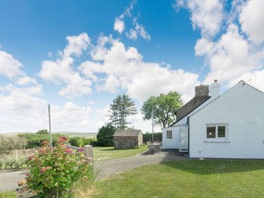 A white house with a small outdoor table and chairs on a paved area beside a garden and a stone shed in the background at Morlan Cottage in Newport Pembrokeshire
