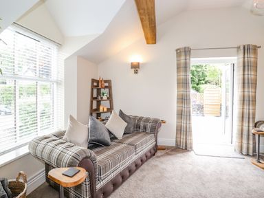 A living room with a plaid sofa and pillows a wooden shelf with decor and candles and a door with plaid curtains at Chestnuts Cottage in Windermere