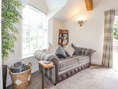 A sitting room with a plaid sofa cushions a basket with blankets a wooden shelf with candles and a window with blinds and curtains at Chestnuts Cottage in Windermere