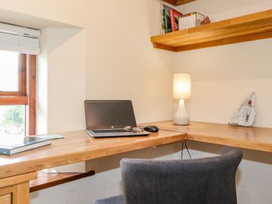 A corner wooden desk with a laptop, glasses, mouse, lamp, books, and a gray chair at Chestnuts Cottage in Windermere