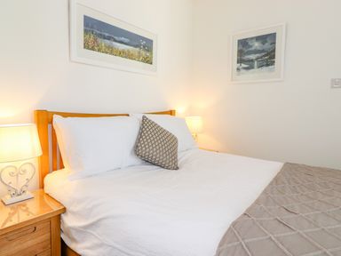 A bedroom with a wooden bed white bedding a patterned cushion two bedside tables with lamps and two pictures on the wall at Chestnuts Cottage in Windermere