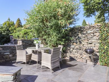 An outdoor patio with a round glass table and four wicker chairs next to a stone wall and a charcoal grill at Chestnuts Cottage in Windermere