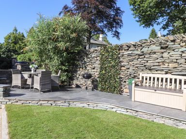 An outdoor patio area with a stone wall wicker chairs a table a barbecue and a bench at Chestnuts Cottage in Windermere