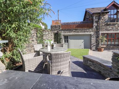 An outdoor patio area with a round glass table and four wicker chairs next to a stone wall and a stone house at Chestnuts Cottage in Windermere