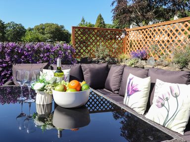 An outdoor seating area with a glass table holding a bowl of fruit wine bottle flowers and three wine glasses at Chestnuts Cottage in Windermere