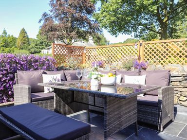 An outdoor patio with wicker furniture including a glass table benches cushions flowers and a wooden lattice fence at Chestnuts Cottage in Windermere