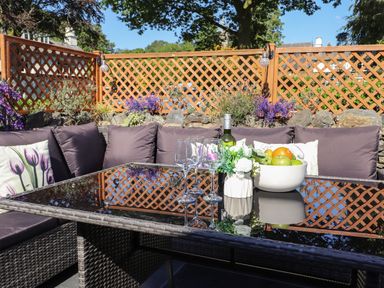 An outdoor patio area with a glass-topped table surrounded by cushioned wicker seating and a wooden lattice fence in the background at Chestnuts Cottage in Windermere
