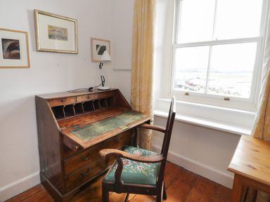 A wooden desk with a green leather writing surface and a chair with a floral cushion near a window with yellow curtains at Wenallt in Porthmadog
