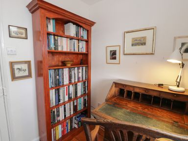 A wooden bookshelf filled with books next to a wooden desk with a lamp and framed pictures on the wall at Wenallt in Porthmadog