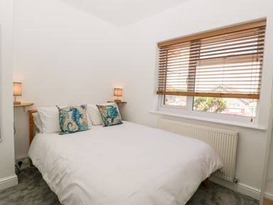 A bedroom with a bed with white bedding and sea animal cushions two wall lamps wooden blinds on window and a radiator below the window at Seacider in Burnham-On-Sea
