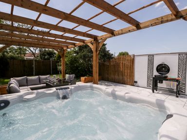 An outdoor patio area with a hot tub under a wooden pergola and a seating area next to a grill at Seacider in Burnham-On-Sea