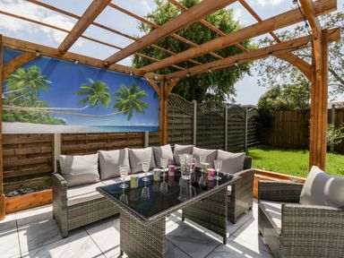 An outdoor seating area with wicker furniture a glass table and a wooden pergola with a tropical beach backdrop at Seacider in Burnham-On-Sea