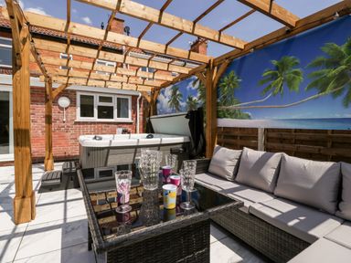 An outdoor patio with a wicker sofa, glass table with cups and glasses, a hot tub under a wooden pergola with a tropical beach mural at Seacider in Burnham-On-Sea
