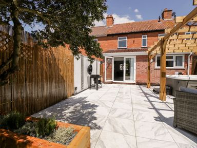 A backyard patio with marble tile flooring, a wooden pergola, outdoor grill, wooden planter, and a brick house with open glass doors at Seacider in Burnham-On-Sea