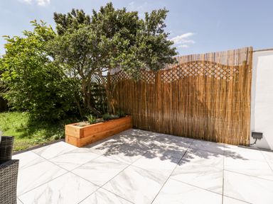 An outdoor patio area with large white marble tiles a wooden planter with a tree and plants a bamboo privacy screen and part of a wicker chair at Seacider in Burnham-On-Sea